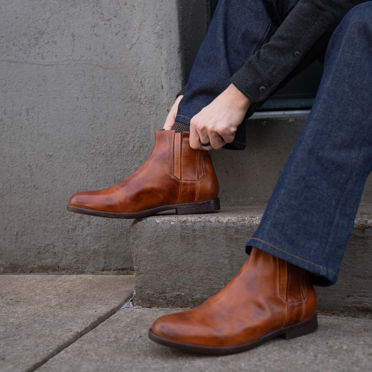 A person in dark jeans and a sweater sits on concrete steps, adjusting the Bed Stu Verrocchio—handcrafted leather slip-on boots.