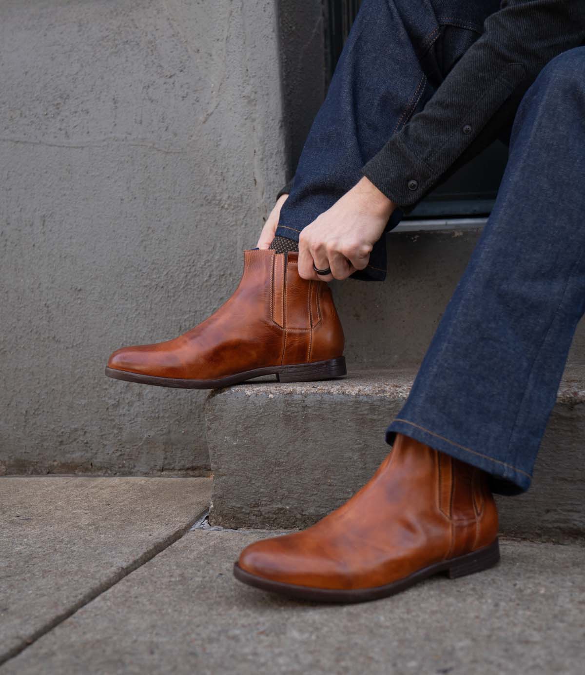 A person in dark jeans and a sweater sits on concrete steps, adjusting the Bed Stu Verrocchio—handcrafted leather slip-on boots.