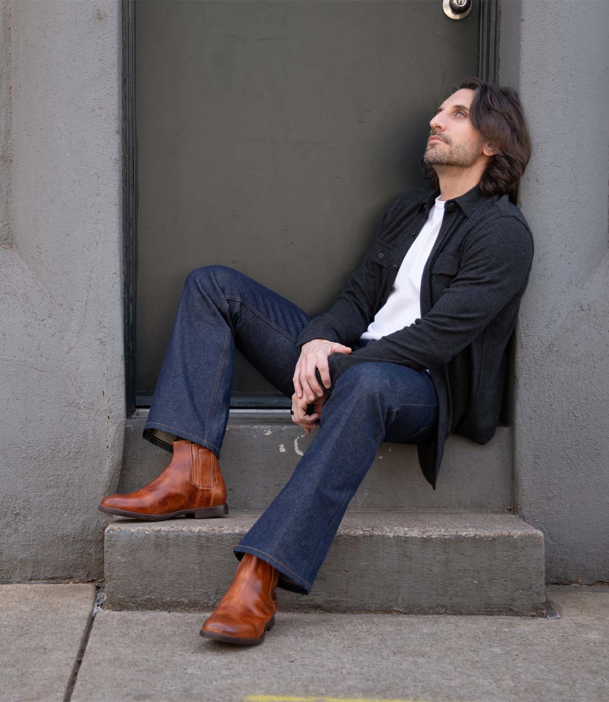 A man with shoulder-length hair sits on a step in front of a door, wearing a dark jacket, white shirt, blue jeans, and Bed Stu Verrocchio handcrafted leather men’s boots, gazing upward.