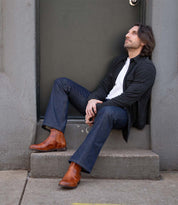 A man with shoulder-length hair sits on a step in front of a door, wearing a dark jacket, white shirt, blue jeans, and Bed Stu Verrocchio handcrafted leather men’s boots, gazing upward.