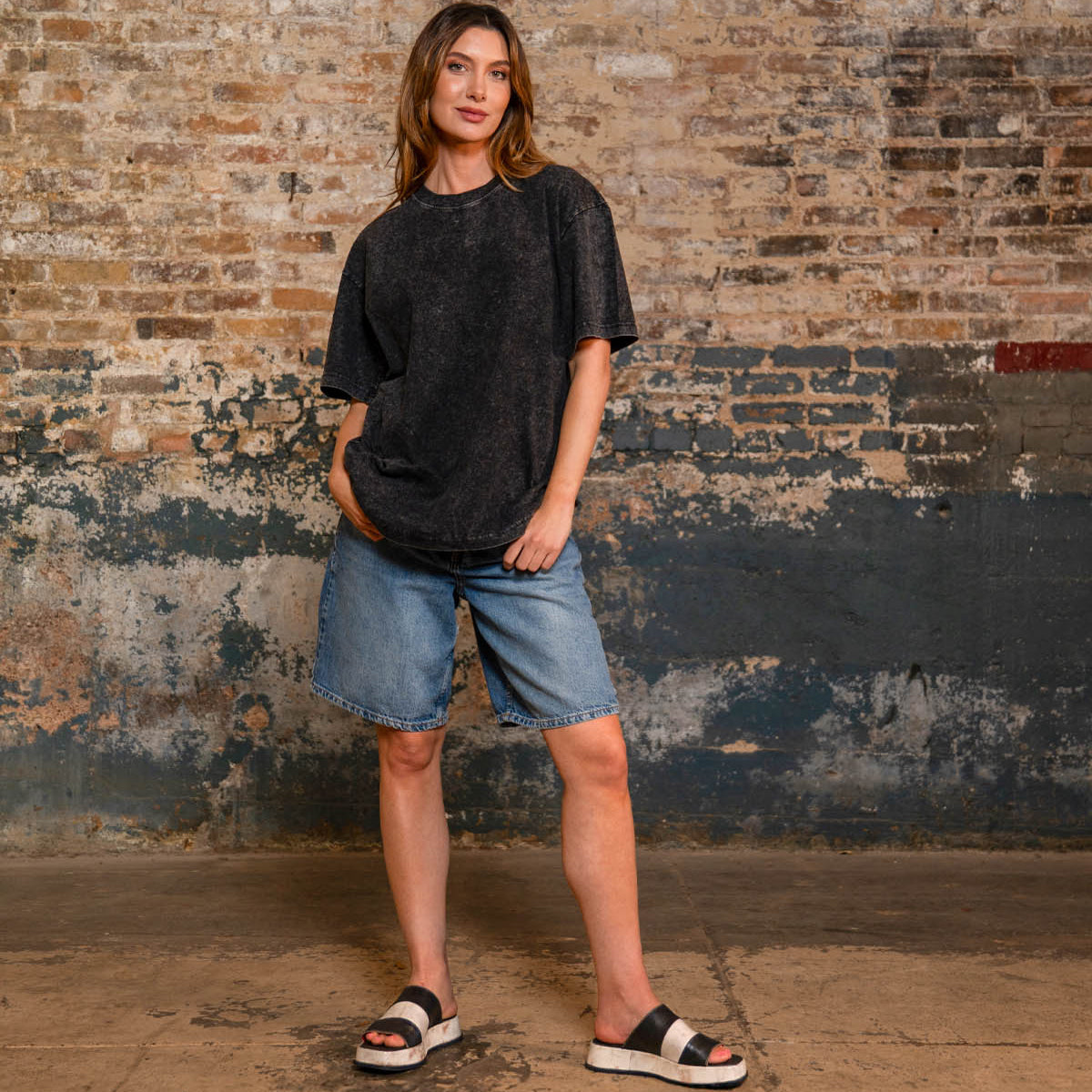 A woman stands indoors by a brick wall, wearing a loose dark tee, denim shorts, and Bed Stu’s Vext platform slide sandals with a leather-wrapped sole.