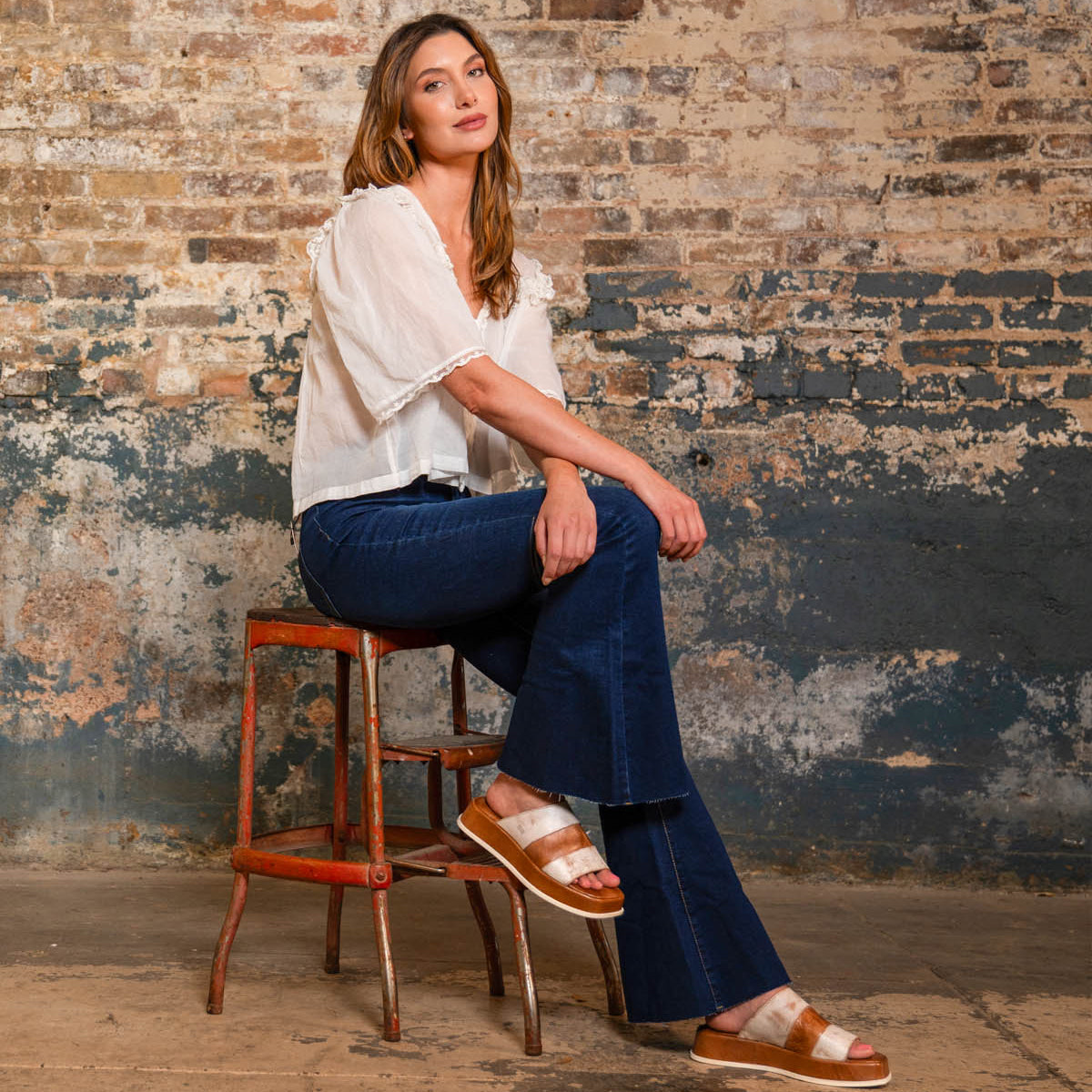 A woman sits on a rustic stool against a weathered brick wall, wearing a white blouse, blue flared jeans, and the Bed Stu Vext platform slide sandals with a leather-wrapped sole.