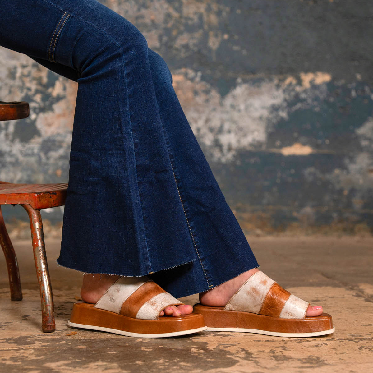A person wearing flared blue jeans and Bed Stu Vext brown and white platform slide sandals with a leather-wrapped sole sits on a rustic metal chair against a textured wall.