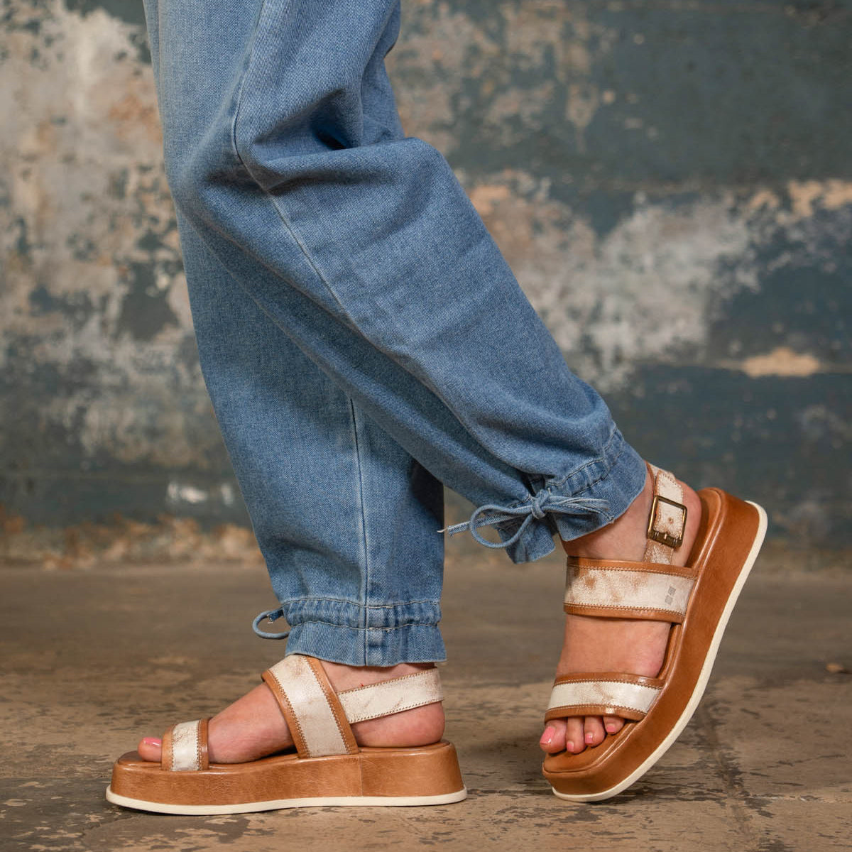 A person wears Bed Stu Vice light brown platform sandals with adjustable ankle strap and light blue jogger jeans, standing on a concrete floor against a distressed wall background.