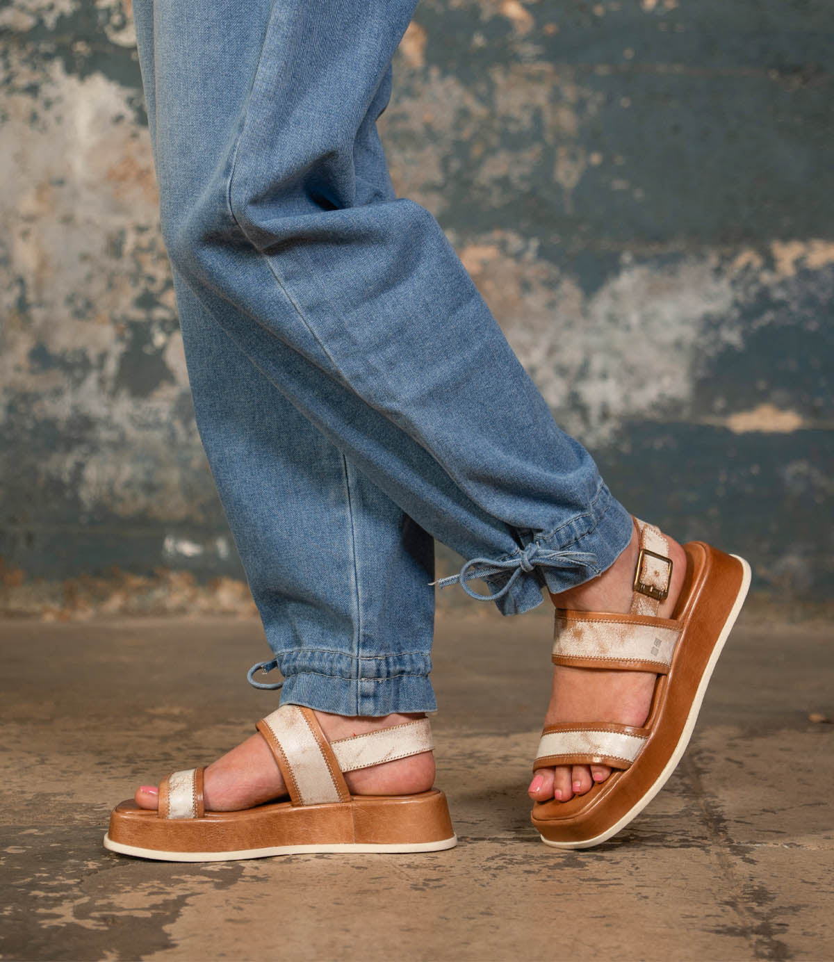 A person wears Bed Stu Vice light brown platform sandals with adjustable ankle strap and light blue jogger jeans, standing on a concrete floor against a distressed wall background.