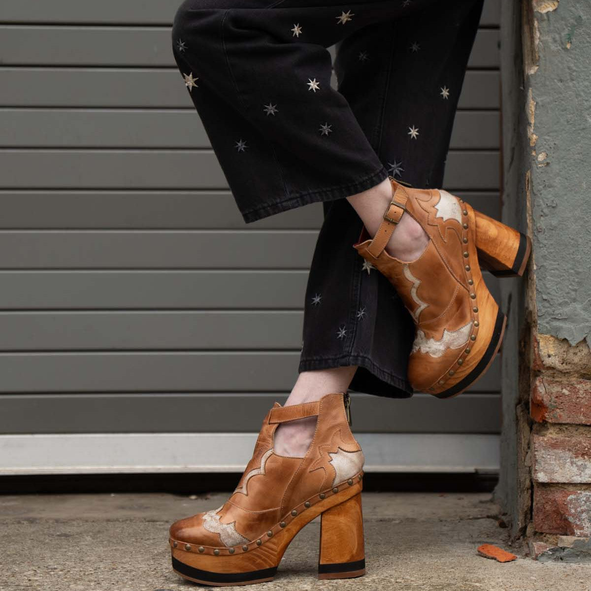A person wearing black star-patterned pants and Bed Stu's Viretta vintage cutout shoes with handcrafted wooden platforms stands by a brick wall.