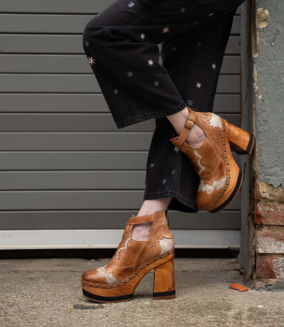 A person wearing black star-patterned pants and Bed Stu's Viretta vintage cutout shoes with handcrafted wooden platforms stands by a brick wall.