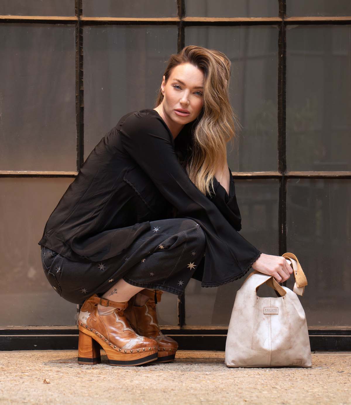 A woman with long blonde hair crouches near a window, wearing a black star-patterned outfit and vintage cutout Bed Stu Viretta tan heeled boots, holding a white handbag.