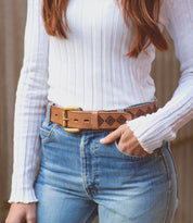 A person is wearing the Wander white ribbed long-sleeve top by Bed Stu, along with blue jeans and a brown belt with whimsical geometric patterns, standing with one hand in the pocket.