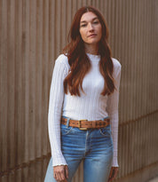 A woman with long red hair, exuding whimsical charm, stands against a wall with vertical grooves while wearing the "Wander" white ribbed sweater by Bed Stu, paired with blue jeans and a brown belt.