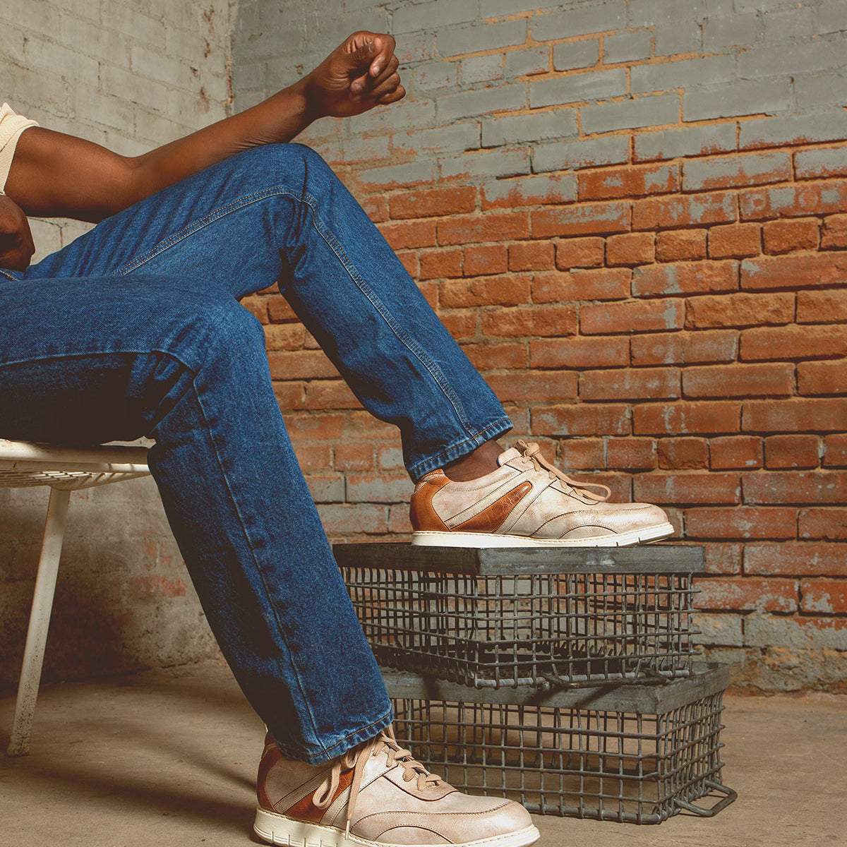 A person sits on a white chair, wearing blue jeans and Wardell lace-up sneakers by Bed Stu. Their foot rests casually on metal crates against a brick wall backdrop.