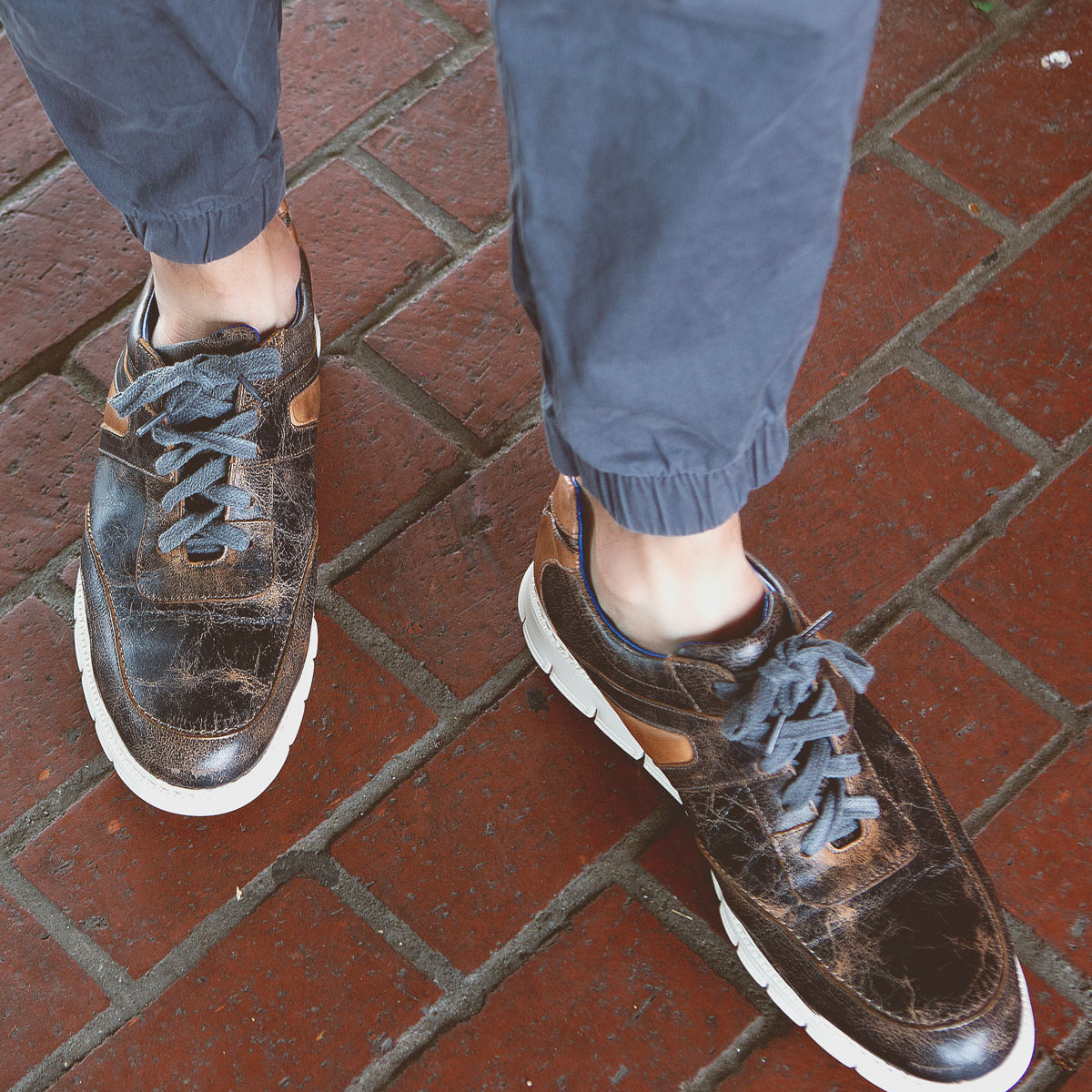 Person wearing navy blue joggers and distressed Bed Stu Wardell lace-up sneakers with white soles, standing on a red brick pavement.
