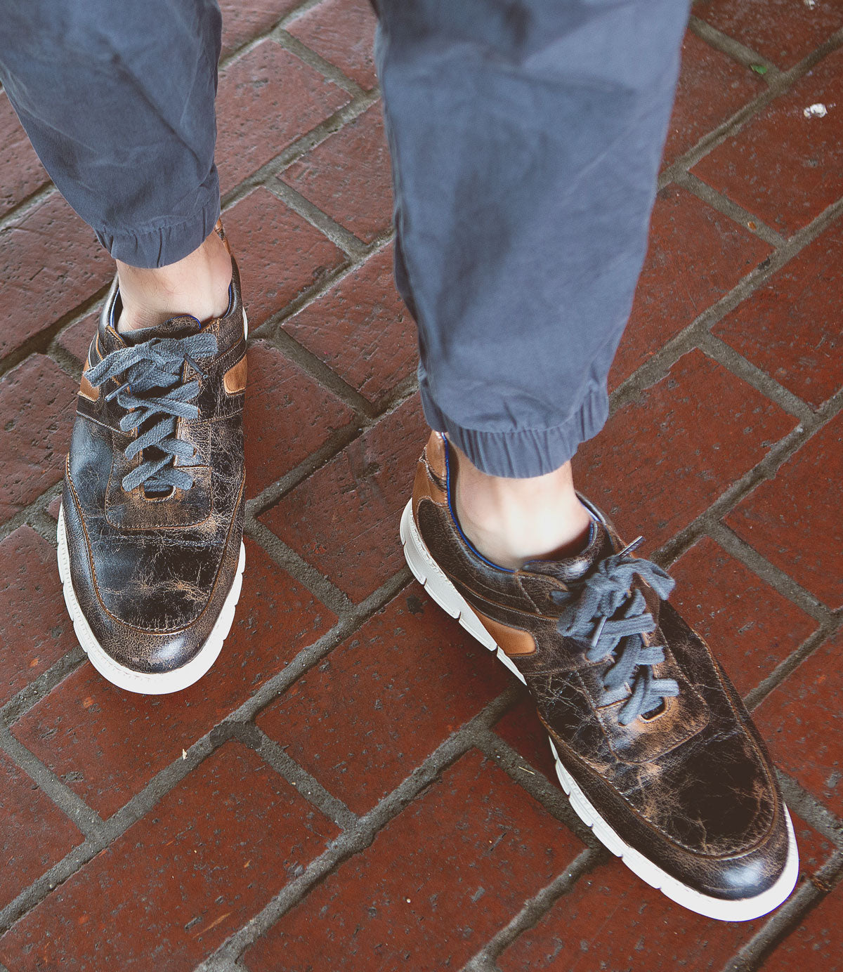 Person wearing navy blue joggers and distressed Bed Stu Wardell lace-up sneakers with white soles, standing on a red brick pavement.