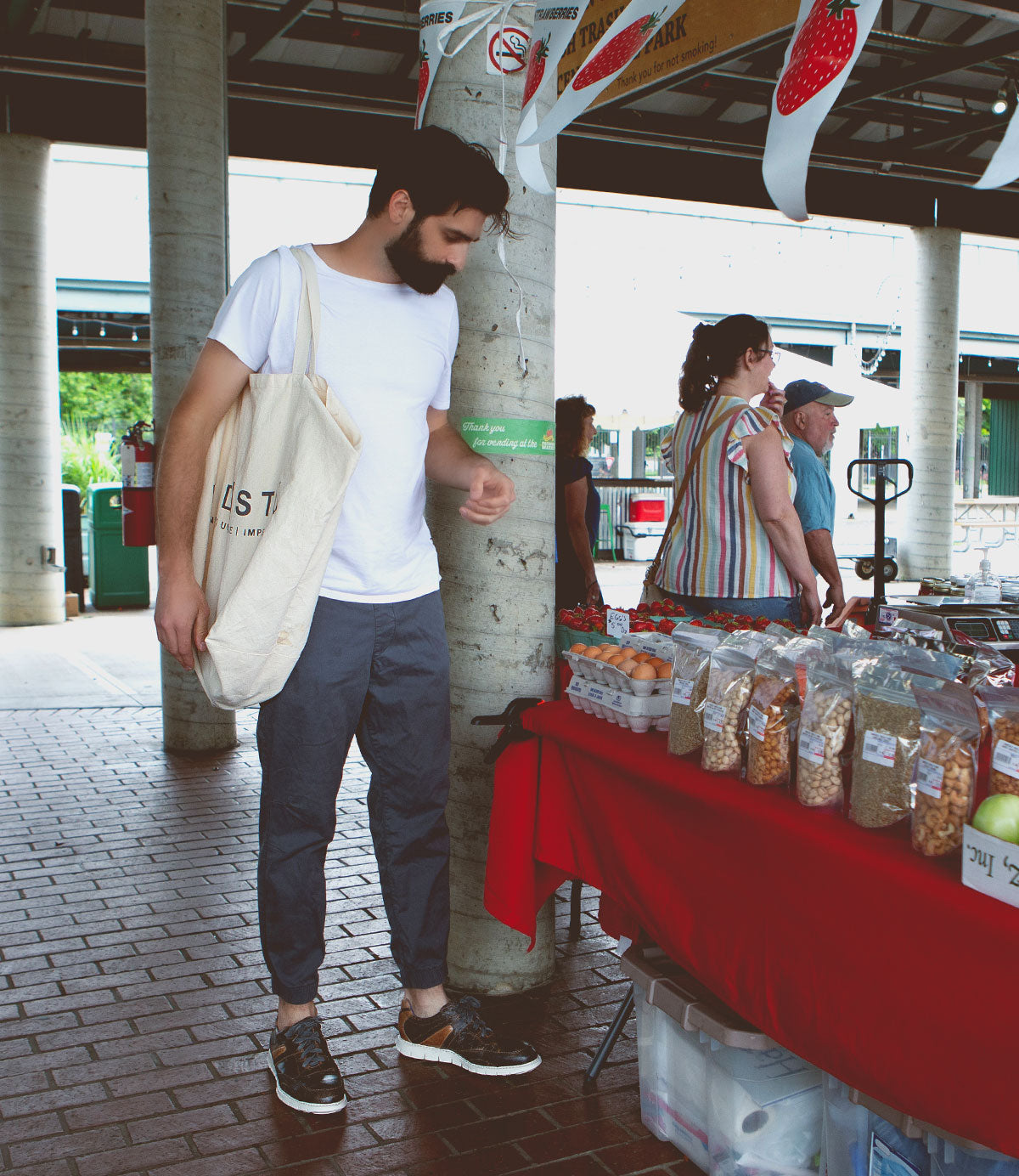 A man with a beard, wearing a white t-shirt and gray pants, looks at items on a market stall table covered with a red cloth. He carries a canvas tote bag over his shoulder and sports distressed Bed Stu Wardell lace-up sneakers. Other people are in the background.