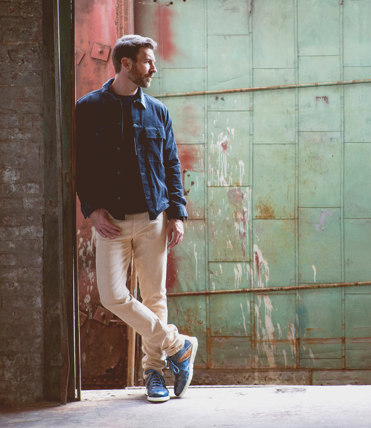 A man in a denim jacket and beige pants leaning against a rustic metal door with a contemplative expression, wearing Bed Stu Wardell distressed lace-up sneakers.