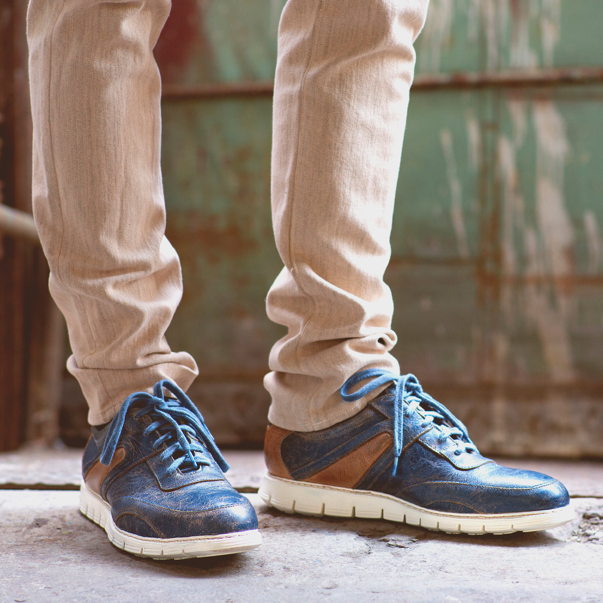 A person standing in an urban setting, wearing distressed Bed Stu lace-up sneakers and beige pants, with a rusted green door in the background.