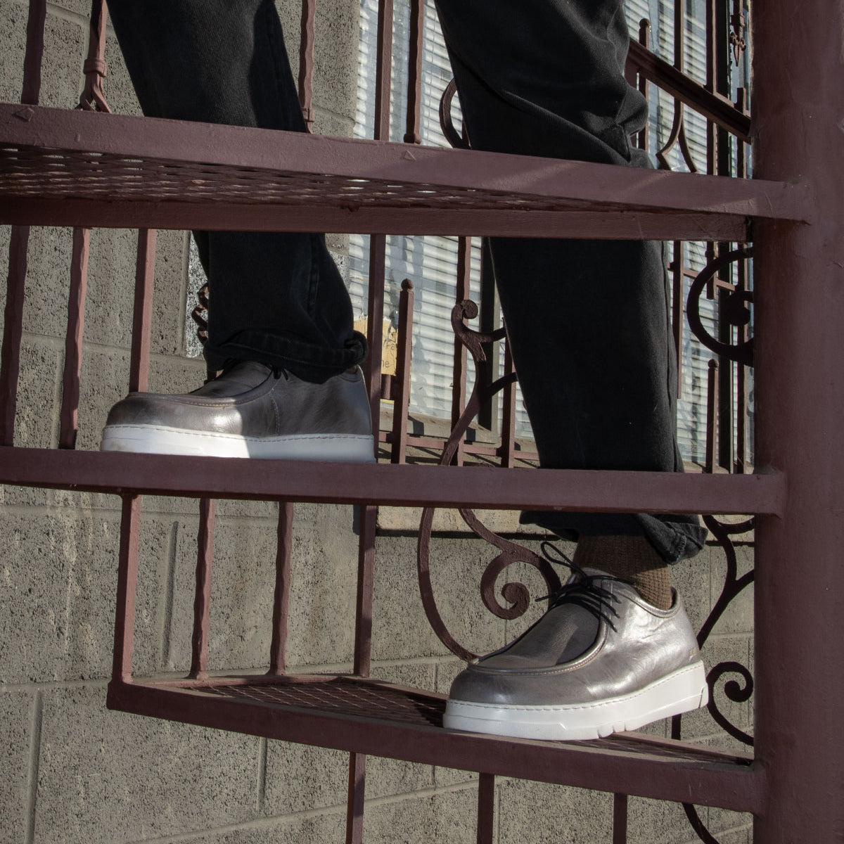 A person wearing the Webmaster slip-on sneakers by Bed Stu, featuring a silver design with white soles, descends a metal spiral staircase. Dressed in black pants, they exude casual style while enjoying all-day comfort.