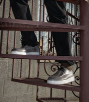 A person wearing the Webmaster slip-on sneakers by Bed Stu, featuring a silver design with white soles, descends a metal spiral staircase. Dressed in black pants, they exude casual style while enjoying all-day comfort.