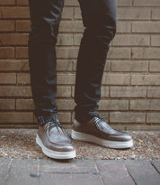 A man in casual style stands in front of a brick wall, wearing Bed Stu's Webmaster shoes with white soles and black pants, exuding an aura of all-day comfort.