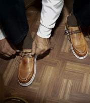 A man ties his Bed Stu Webmaster lace-up moccasins on a wooden floor, savoring their all-day comfort.