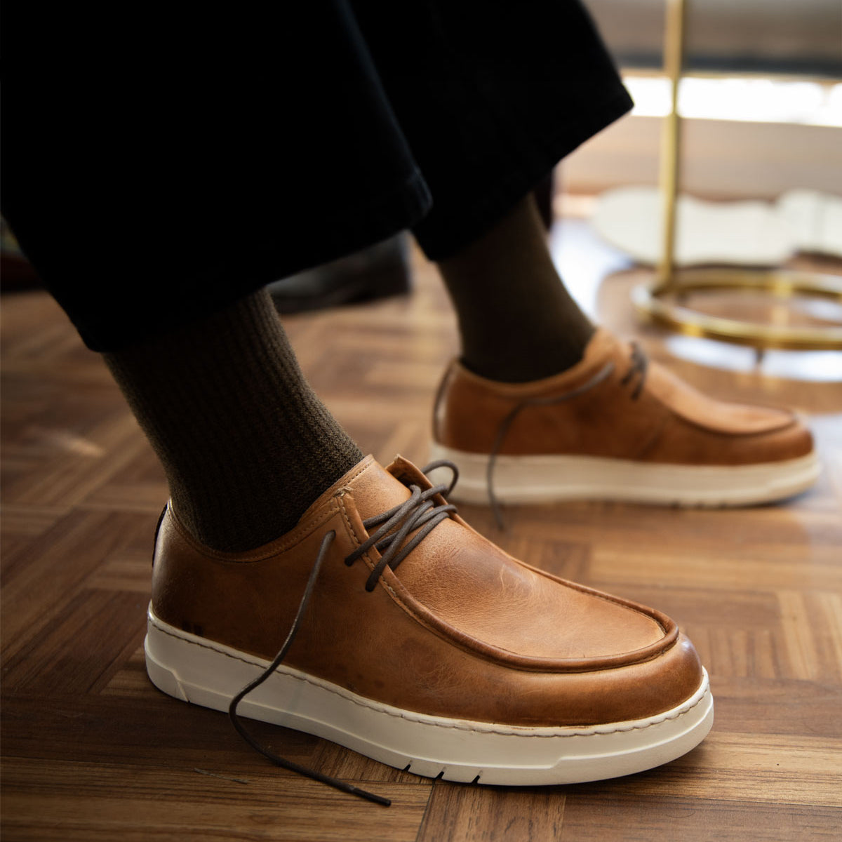 The Bed Stu Webmaster sneakers, featuring brown leather with white soles, exude casual style on a wooden floor. Perfect for pairing with dark pants and socks, they promise all-day comfort.