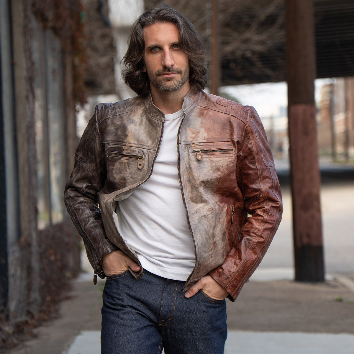 A man with shoulder-length hair wears the Bed Stu Windfall brown leather jacket with satinized cotton lining, paired with a white t-shirt and jeans, standing outdoors on a city sidewalk.