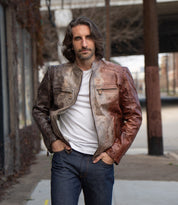 A man with shoulder-length hair wears the Bed Stu Windfall brown leather jacket with satinized cotton lining, paired with a white t-shirt and jeans, standing outdoors on a city sidewalk.