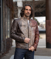 A man with shoulder-length hair stands on a city sidewalk wearing Bed Stu's Windfall leather jacket, which features two-toned brown and reddish hues, satinized cotton lining, and is paired with blue jeans.