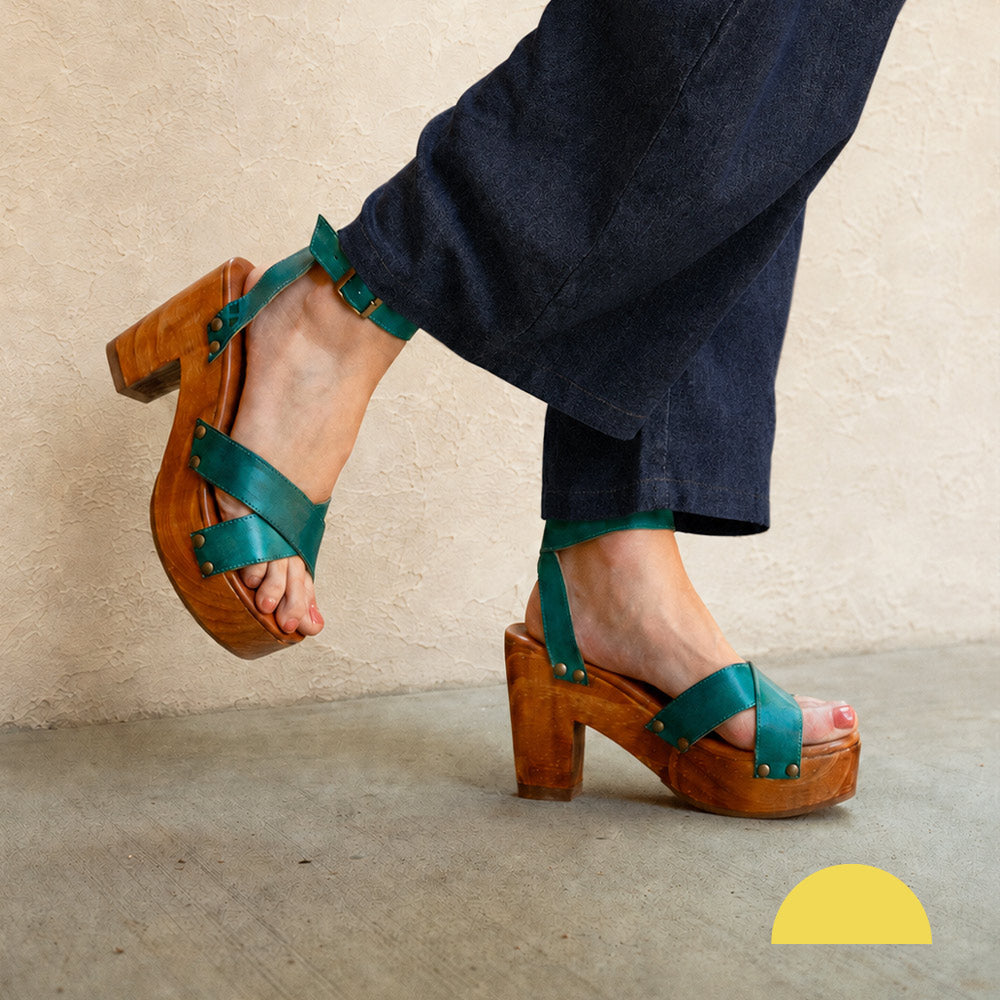 Person wearing teal strappy platform sandals with chunky wooden heels and dark cropped pants.