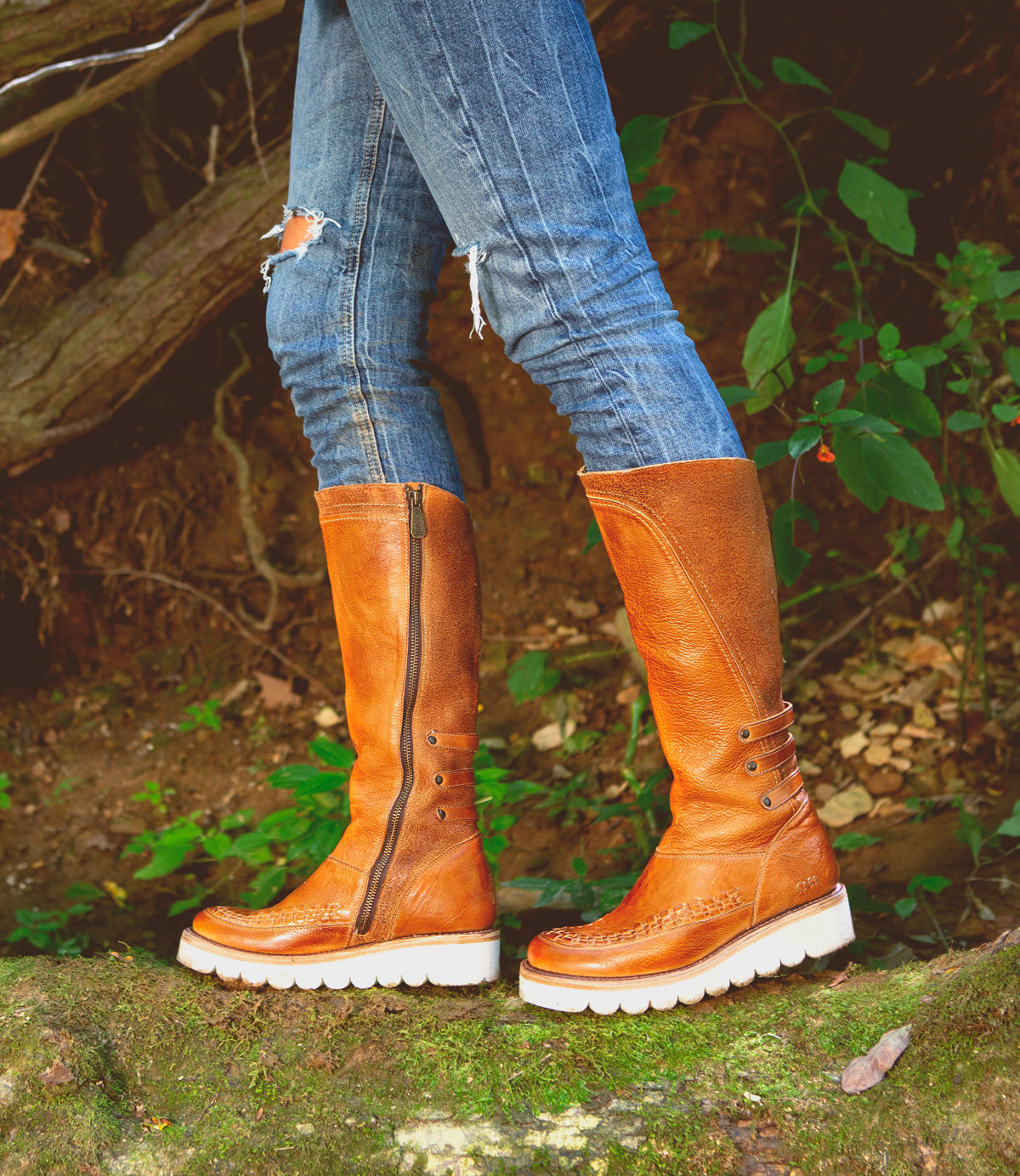 A woman in tall Bed Stu leather boots standing on a log in the woods.