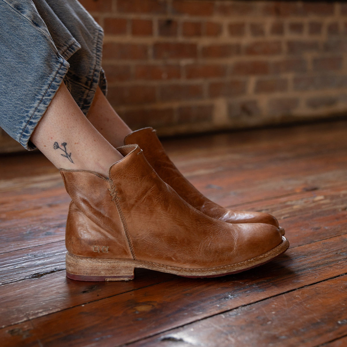 A person wears Bed Stu Yurisa light brown leather ankle boots with rolled-up jeans, sitting on a wooden floor by a brick wall. The everyday boot features a red leather lining and reveals a small flower tattoo on the ankle.