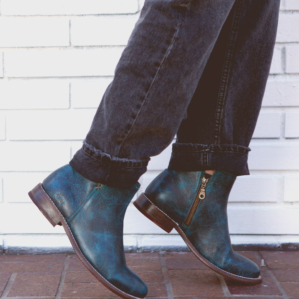 A person wearing Yurisa dark blue leather ankle boots with a side zipper by Bed Stu and black jeans stands on a brick pavement against a white brick wall, showcasing the timeless design and practical elegance of an everyday boot.