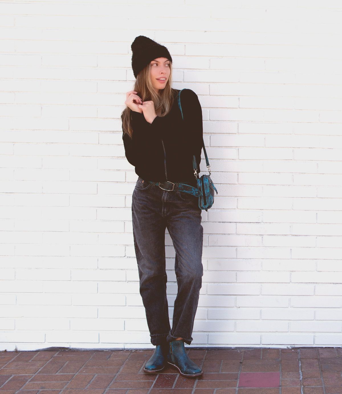 A person stands against a white brick wall, sporting a black hat, black long-sleeve top, dark jeans, and Bed Stu's Yurisa everyday teal boots with a timeless design. A matching teal handbag hangs from their shoulder.