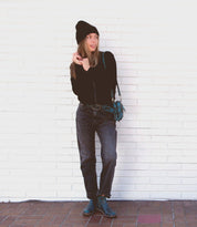 A person stands against a white brick wall, sporting a black hat, black long-sleeve top, dark jeans, and Bed Stu's Yurisa everyday teal boots with a timeless design. A matching teal handbag hangs from their shoulder.
