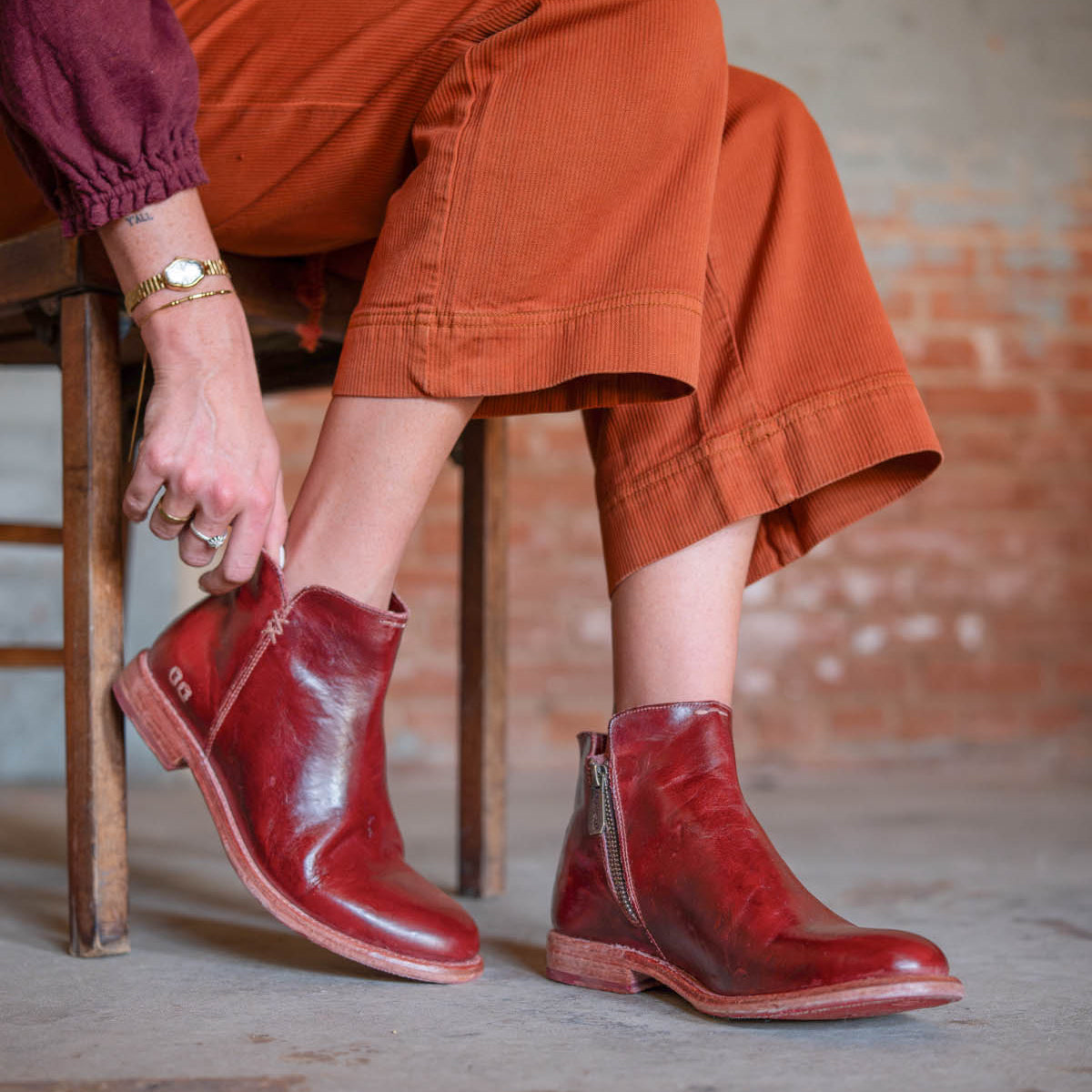 A person in rust-colored pants sits on a wooden chair, adjusting one Yurisa boot by Bed Stu—red leather ankle boots featuring side zippers, a Vibram outsole, and red leather lining.