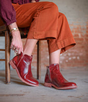 A person in rust-colored pants sits on a wooden chair, adjusting one Yurisa boot by Bed Stu—red leather ankle boots featuring side zippers, a Vibram outsole, and red leather lining.