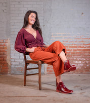 A woman with long brown hair sits on a wooden chair, wearing the Bed Stu Yurisa red ankle boots with a Vibram outsole, maroon blouse, and orange pants, set against a brick and gray painted wall.
