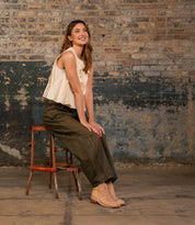 A woman sits sideways on a stool against a distressed brick wall, wearing a sleeveless beige top, olive green pants, and Bed Stu Yurisa tan everyday boots with a Vibram outsole.