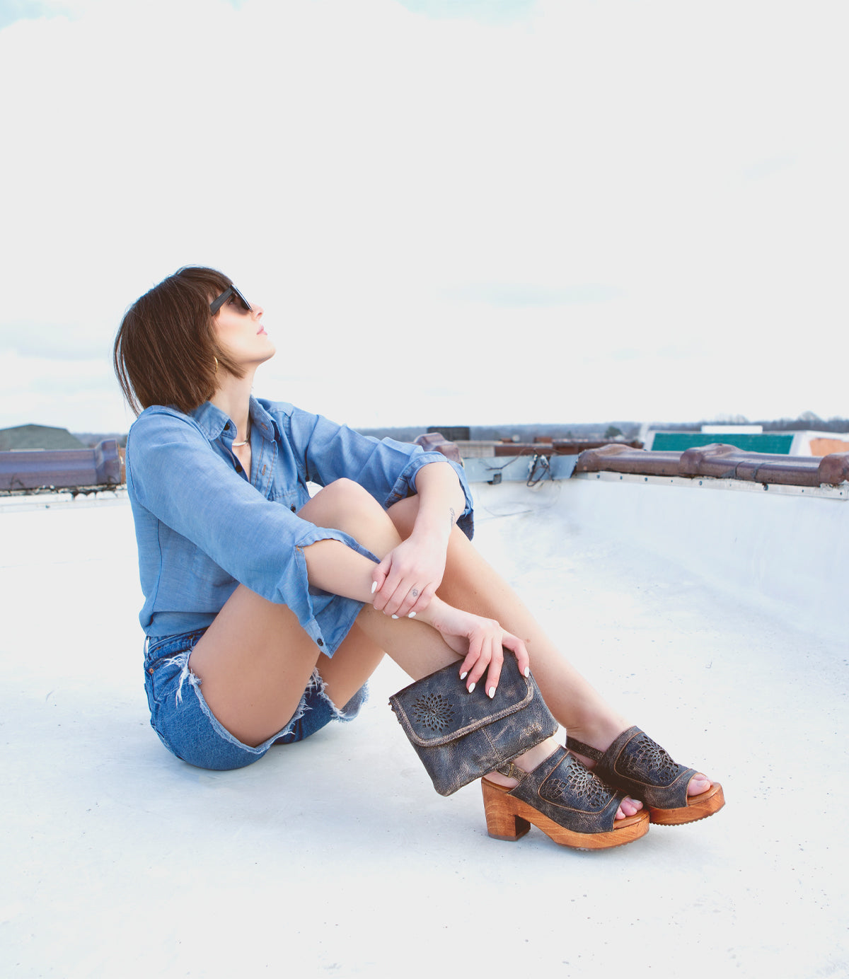 A woman sitting on top of a roof wearing a denim shirt and sandals, while showcasing her stylish accessory - the Bed Stu Ziggy II leather crossbody bag with multiple compartments.