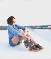 A woman sitting on top of a roof wearing a denim shirt and sandals, while showcasing her stylish accessory - the Bed Stu Ziggy II leather crossbody bag with multiple compartments.