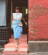 A woman wearing a blue midi skirt and white top, accessorized with a stylish Bed Stu Ziggy II leather crossbody bag with compartments.