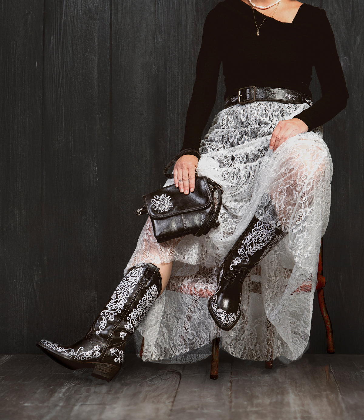 A person sits on a chair in a black top, white lace skirt, and Bed Stu Ziggy JB embroidered black cowboy boots, holding a sleek black crossbody clutch against a dark wooden backdrop.