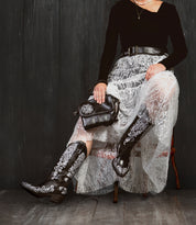 A person sits on a chair in a black top, white lace skirt, and Bed Stu Ziggy JB embroidered black cowboy boots, holding a sleek black crossbody clutch against a dark wooden backdrop.
