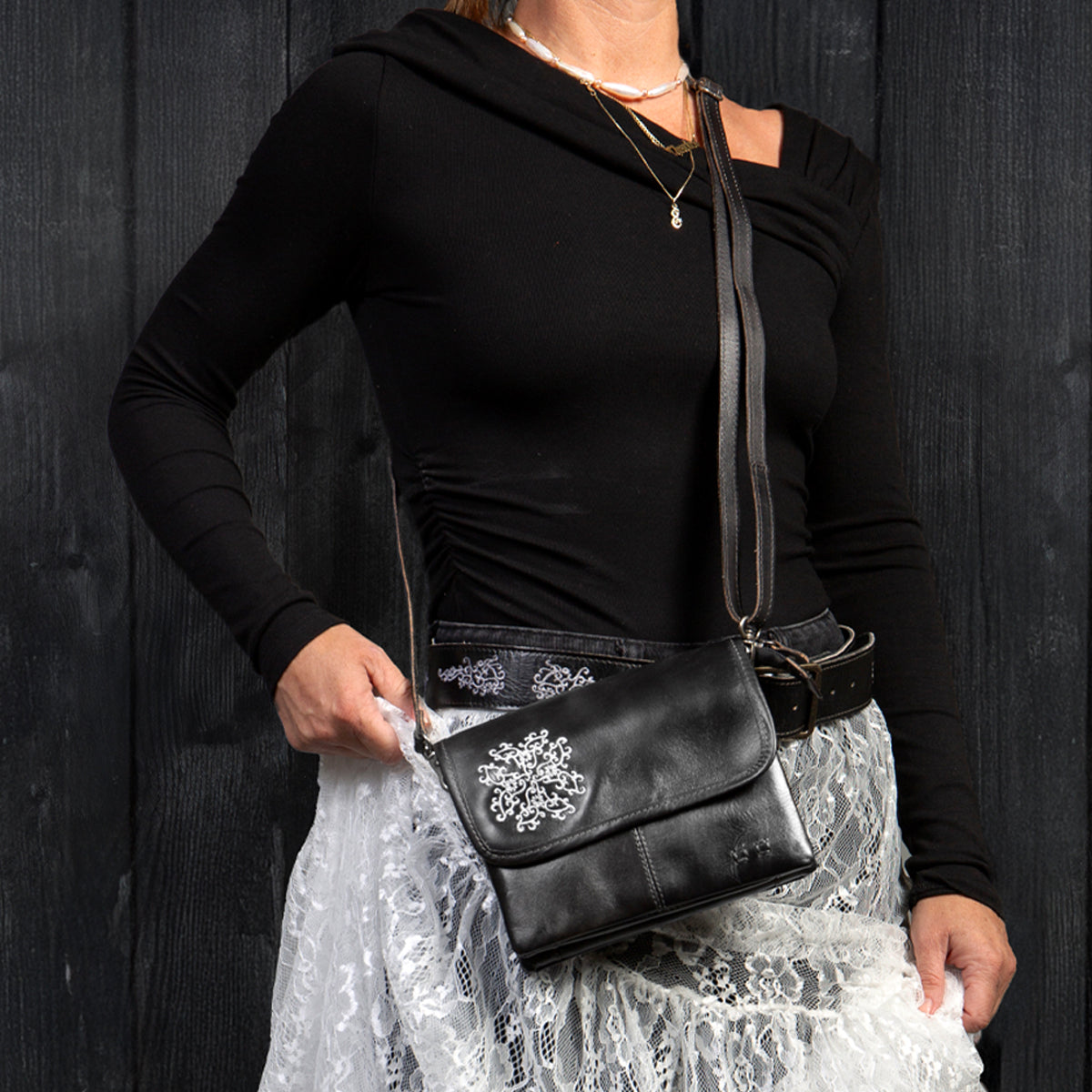 A person in a black asymmetrical top and white lace skirt holds the Bed Stu Ziggy JB crossbody clutch with a silver floral design against a dark wooden background.