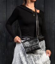 A person in a black asymmetrical top and white lace skirt holds the Bed Stu Ziggy JB crossbody clutch with a silver floral design against a dark wooden background.