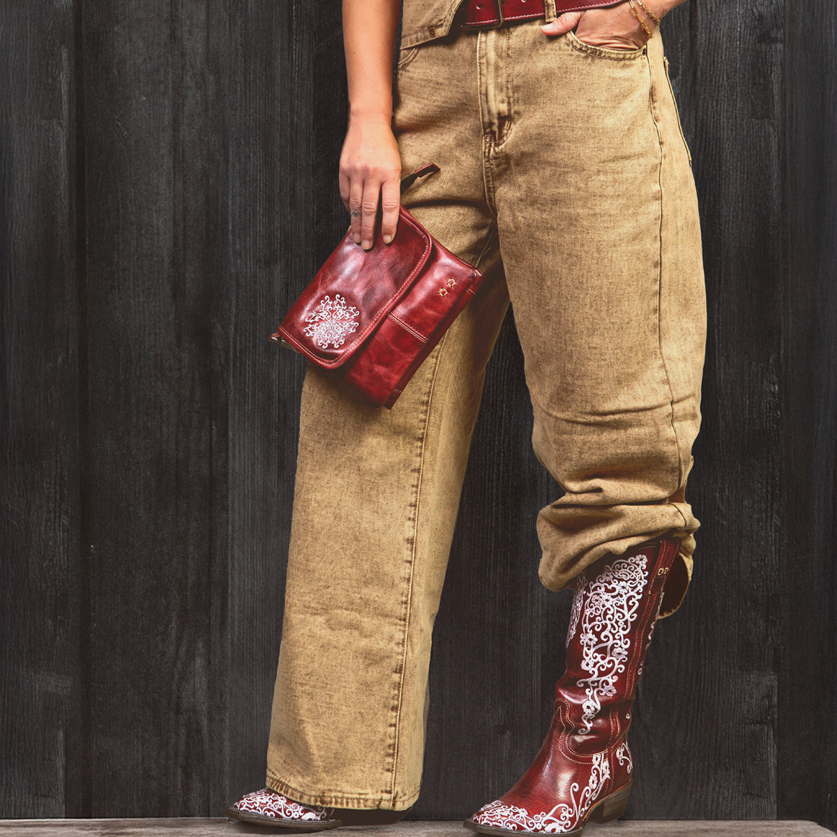 A person stands against a dark wooden wall wearing Bed Stu’s Ziggy JB tan overalls with a red belt, matching red fold-over flap bag, and red cowboy boots detailed with white embroidery.