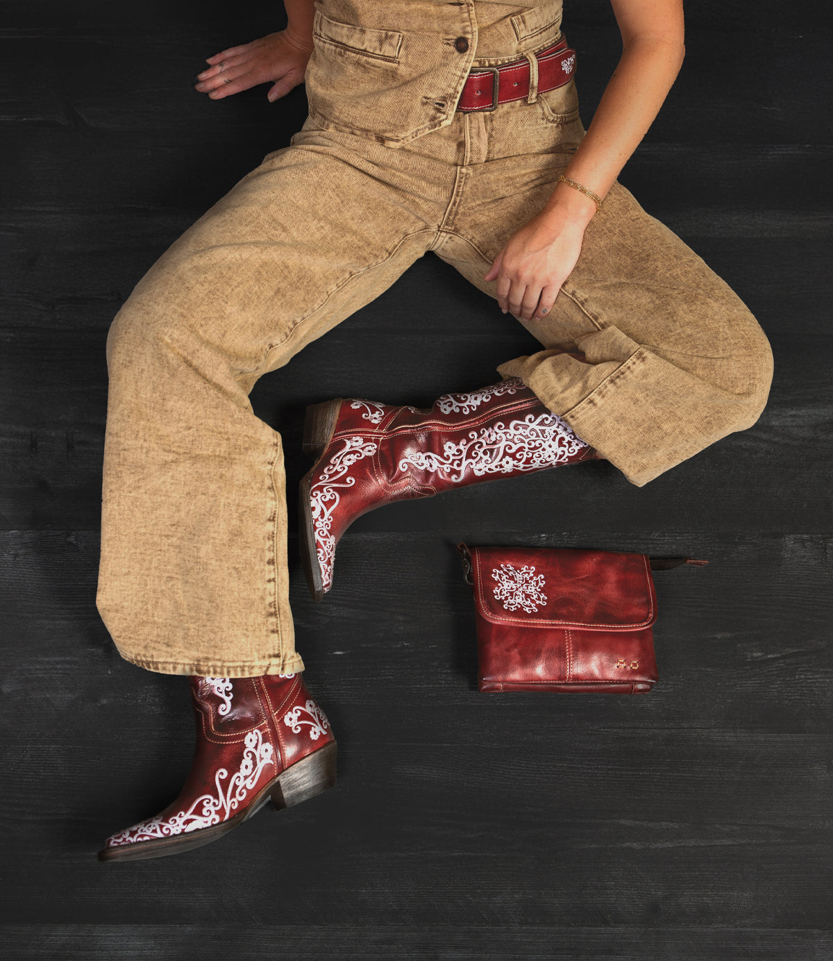 A person wearing tan overalls with red and white Bed Stu Ziggy JB western boots sits on a black surface beside a matching red leather fold-over flap bag.