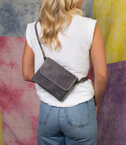 A person with blonde hair, in a white sleeveless top and blue jeans, stands with their back to the camera wearing the Bed Stu Ziggy gray leather crossbody bag against a colorful fabric background.