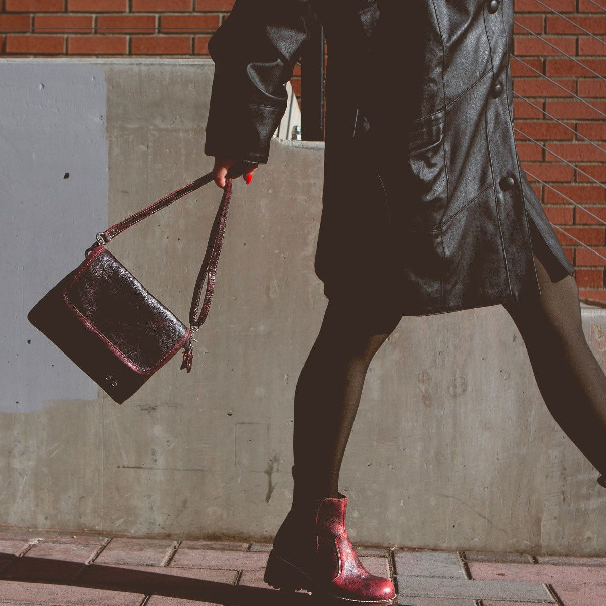 A person in a black leather coat, black leggings, and a pair of Bed Stu's Ziggy red boots strides along a sidewalk, holding a trendy belt bag with a brick wall in the background.