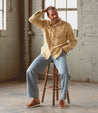 A man in a beige shirt and blue jeans sitting on a wooden stool, smiling and posing with one hand on his head and wearing Bed Stu leather sandals in a bright, industrial-style room.